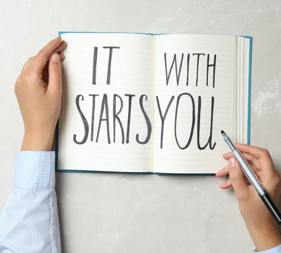 Picture of a business man's hand writing in a journal, the pages read, 'It Starts With You' referring to the first part of working with a VA which begins with a chat about your business needs.