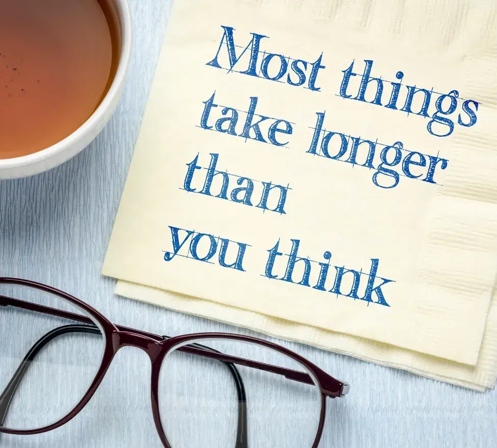 Picture of a napkin next to a pair of reading glasses. Text on the napkin reads: Most things take longer than you think.