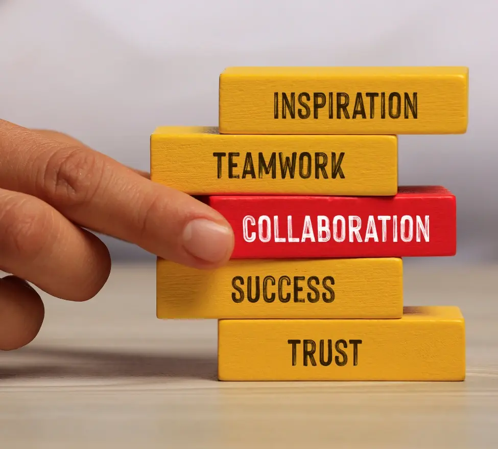 Picture of jenga style blocks on a table being laid, each block has a word: Inspiration, Team work, Collaboration, Success and Trust. They are yellow blocks with the Collaboration block in red., 